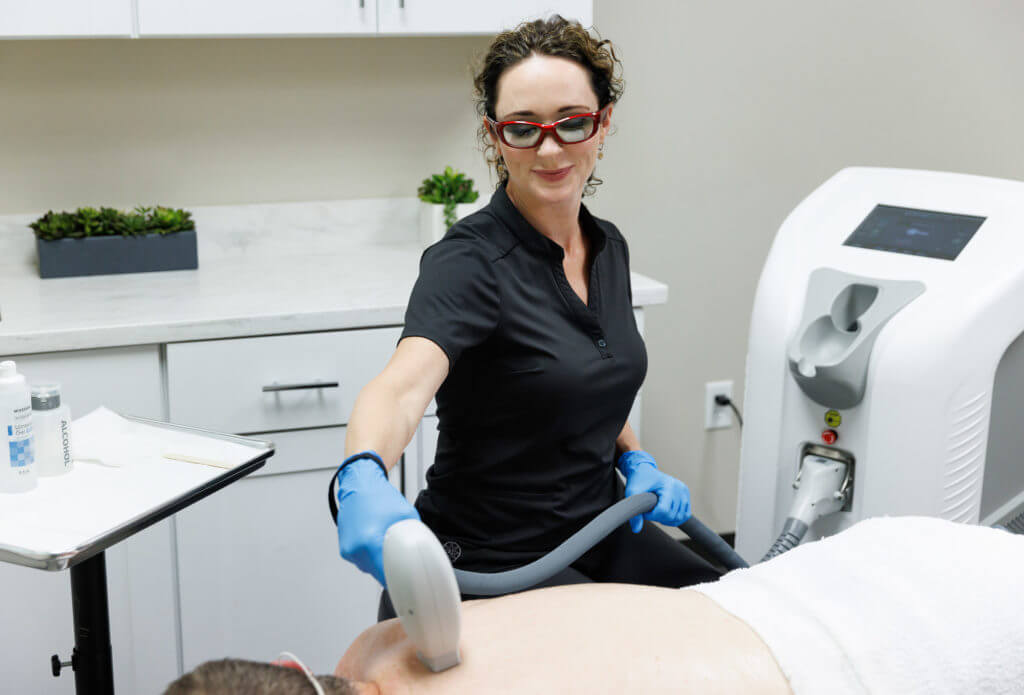 A female clinician wearing protective glasses and blue gloves performs a laser treatment on a patient’s back using a handheld device connected to a large medical laser machine in a clinical setting.