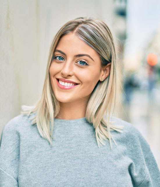 A smiling woman with straight, shoulder-length blonde hair parted in the middle, wearing a light grey top, standing outdoors against a light-colored wall with a blurred city street in the background.