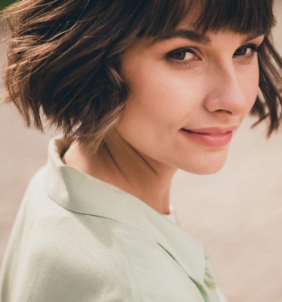 A woman with short, wavy brown hair and bangs wears a light green collared shirt, glancing over her shoulder with a subtle smile in an outdoor setting.