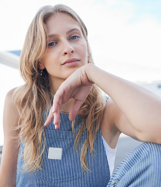 A young woman with long, wavy blonde hair wears a blue-and-white striped overall dress over a white top, resting her chin on her hand and gazing calmly at the camera in an outdoor setting.