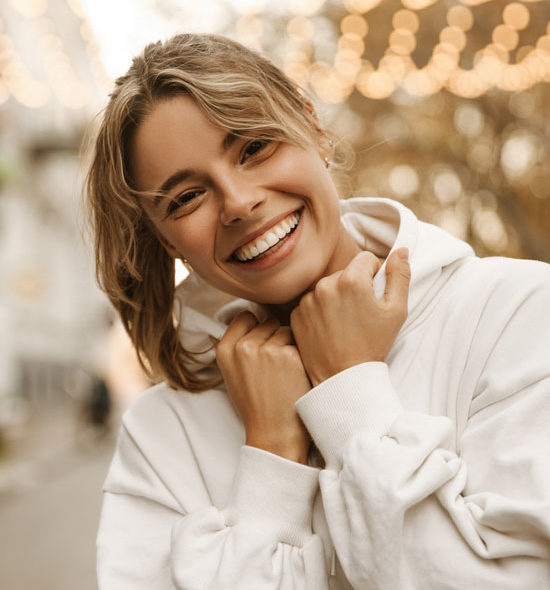 A young woman with light brown hair tied back and loose strands framing her face smiles brightly, wearing a white hoodie and holding its collar, standing outdoors with warm bokeh string lights in the blurred background.