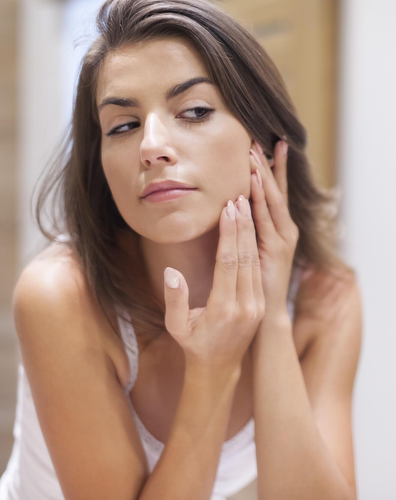 Woman examining her skin in the mirror, gently touching her cheek with a concerned expression.