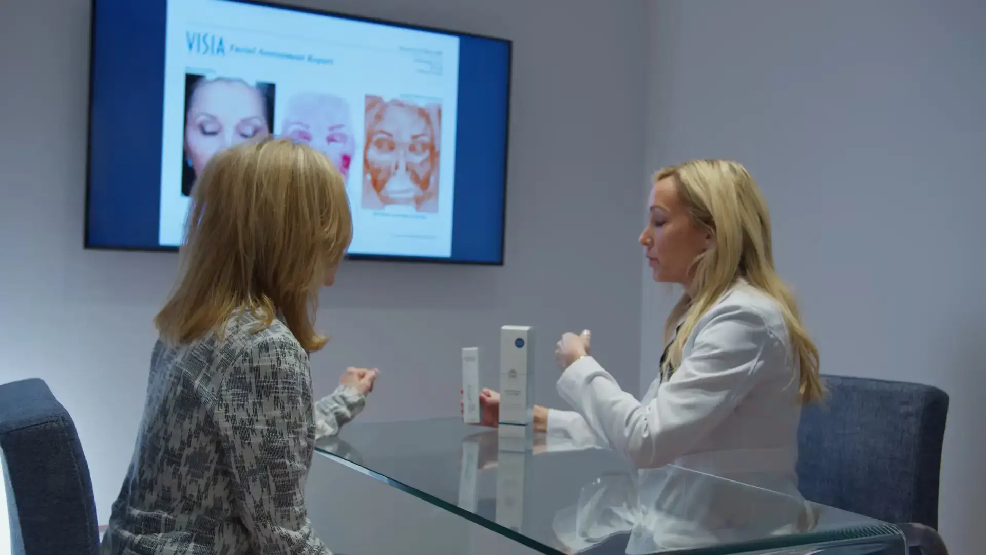 Two women seated at a glass table in a consultation room, discussing skincare products with a presentation on skin treatments displayed on a screen in the background.