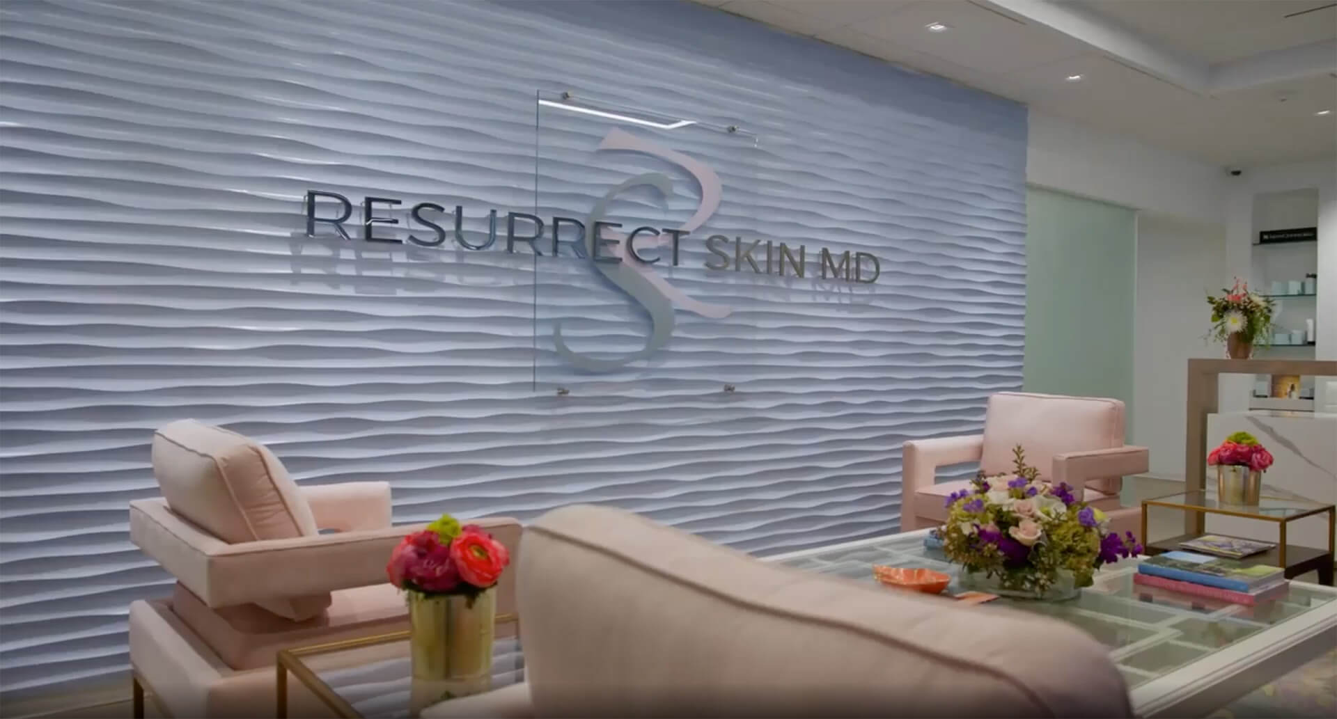 Modern waiting area at Resurrect Skin MD with pink chairs, floral arrangements, a glass coffee table, and a textured wall featuring the clinic’s logo.