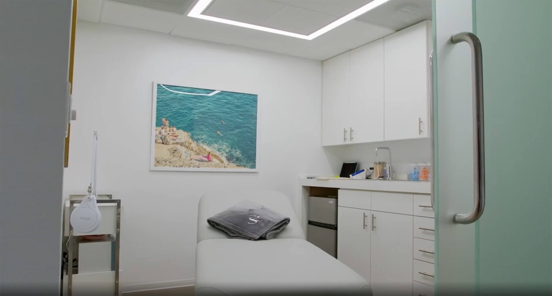 Modern medical treatment room with a patient bed, white cabinetry, small sink area, and ocean-themed wall art creating a clean and calming environment.