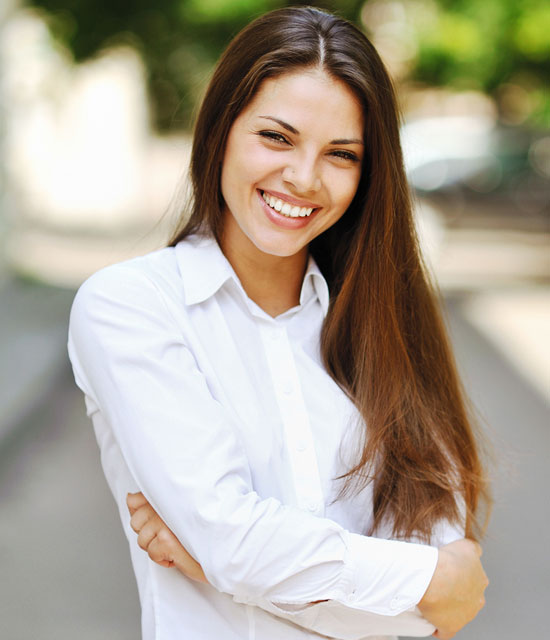 Smiling young woman with long brown hair wearing a white shirt, standing outdoors with arms crossed.