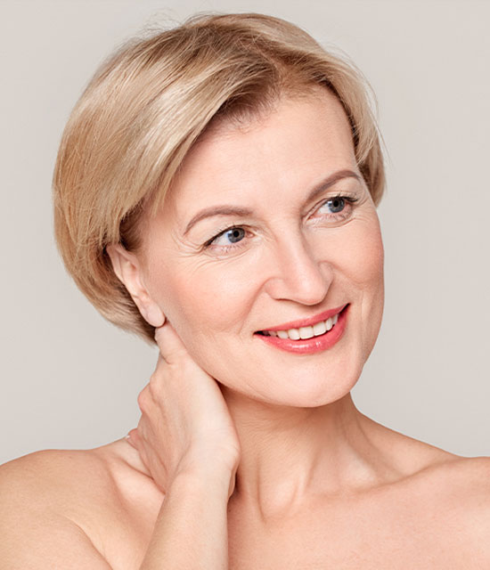 Smiling middle-aged woman with short blonde hair, clear skin, and natural makeup gently touching her neck while looking to the side