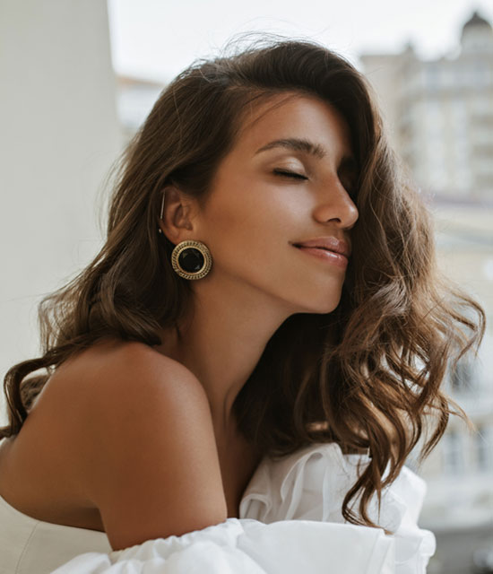 Smiling woman with wavy brown hair, wearing statement earrings and an off-shoulder white top, posing outdoors with eyes closed