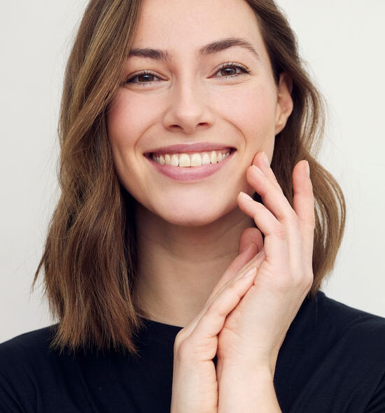 Smiling woman with shoulder-length light brown hair, wearing a black top and gently touching her face with both hands.