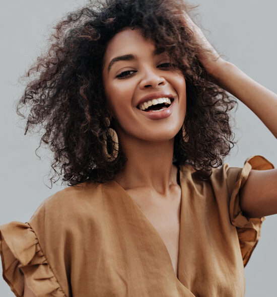 Smiling woman with curly hair wearing a brown blouse and hoop earrings, posing confidently outdoors