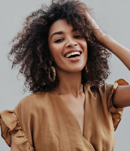 Smiling woman with curly hair wearing a brown blouse and hoop earrings, posing confidently outdoors