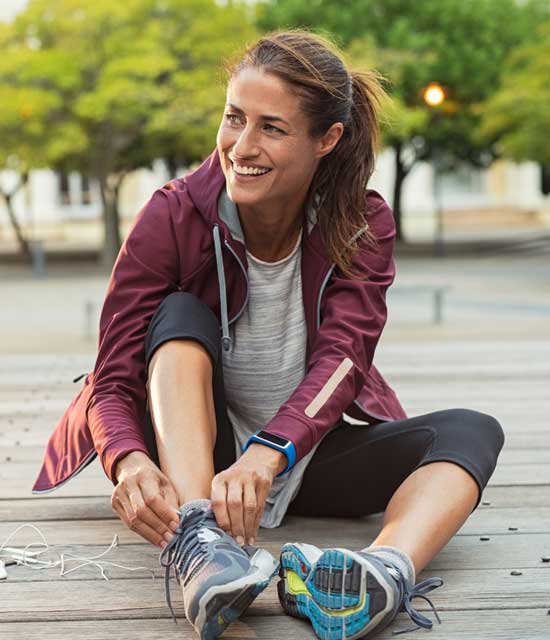 Smiling woman in athletic wear sitting outdoors, tying her running shoes after a workout.