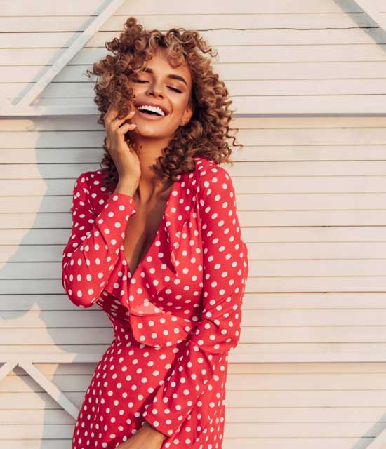 Joyful woman with curly hair wearing a red polka dot dress, smiling brightly while posing outdoors.