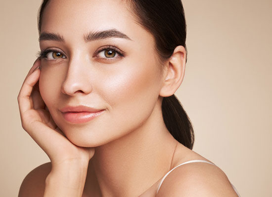 Smiling woman with smooth, radiant skin resting her face on her hand, wearing a white spaghetti strap top against a warm background.