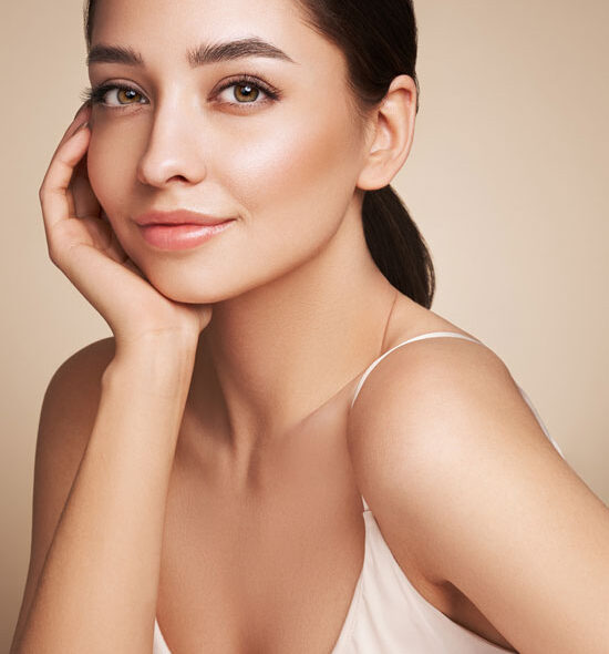 Smiling woman with smooth, radiant skin resting her face on her hand, wearing a white spaghetti strap top against a warm background.
