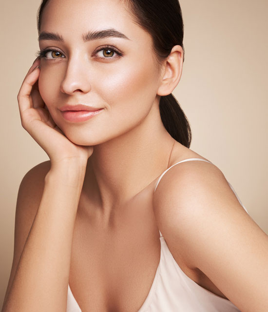 Smiling woman with smooth, radiant skin resting her face on her hand, wearing a white spaghetti strap top against a warm background.