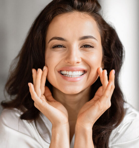 Smiling woman with glowing skin and dark wavy hair touching her face gently, showcasing a fresh and radiant complexion