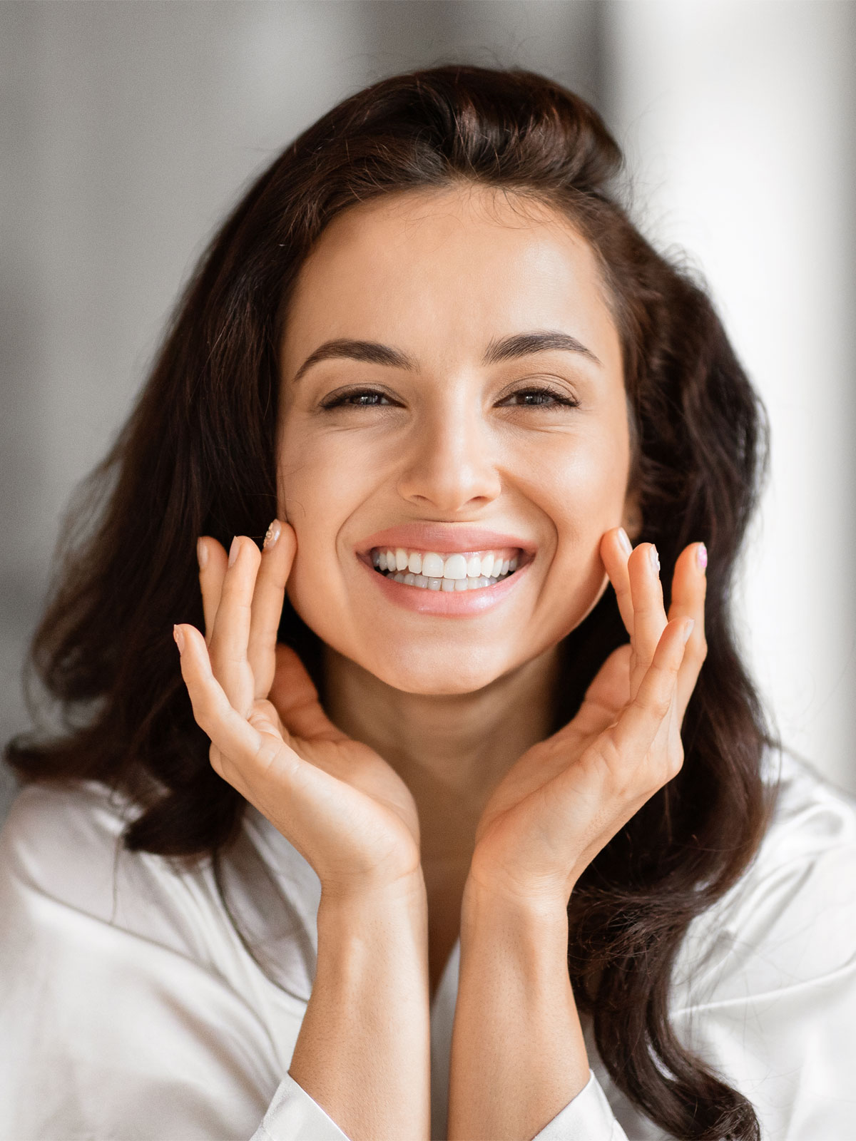 Smiling woman with glowing skin and dark wavy hair touching her face gently, showcasing a fresh and radiant complexion