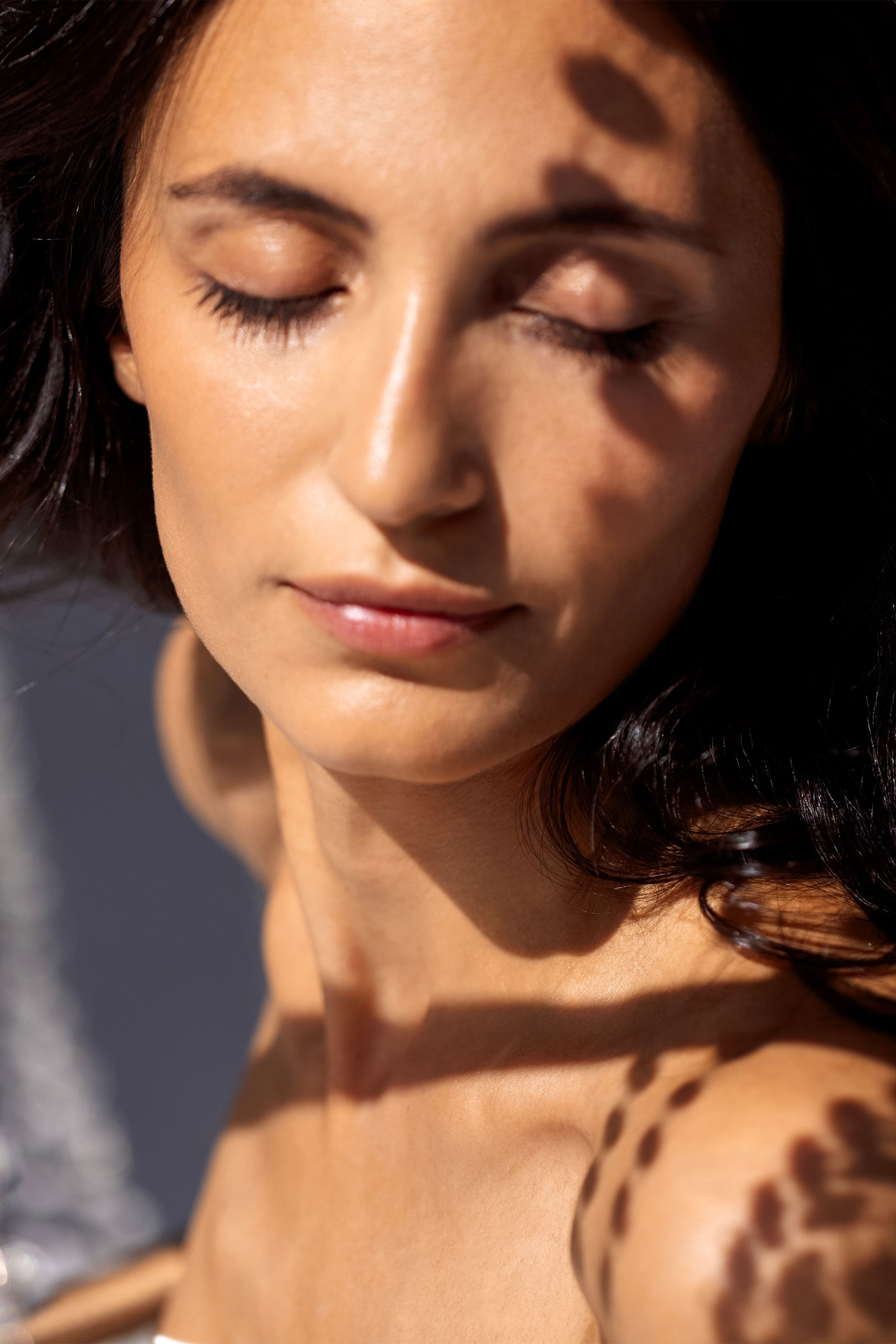 Woman with natural glowing skin and dark hair, eyes closed while sunlight and leaf shadows create patterns across her face and shoulders