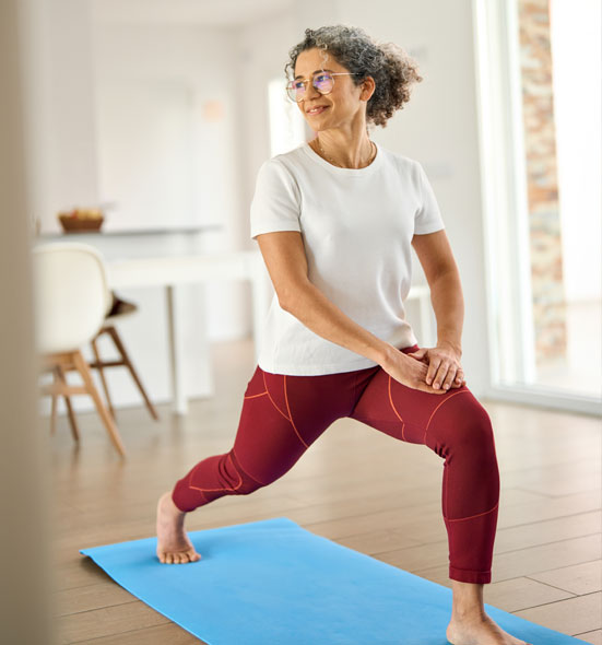 Smiling older woman with curly gray hair doing a lunge stretch on a yoga mat at home, wearing a white shirt and red leggings