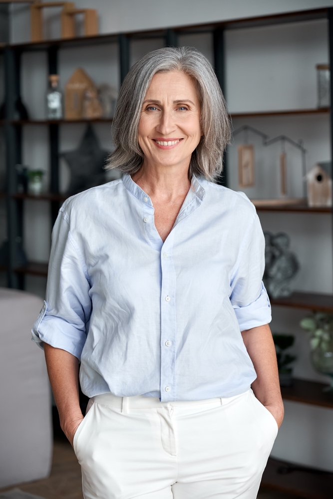 Smiling middle-aged woman with gray hair wearing a light blue blouse and white pants, standing confidently indoors with hands in pockets