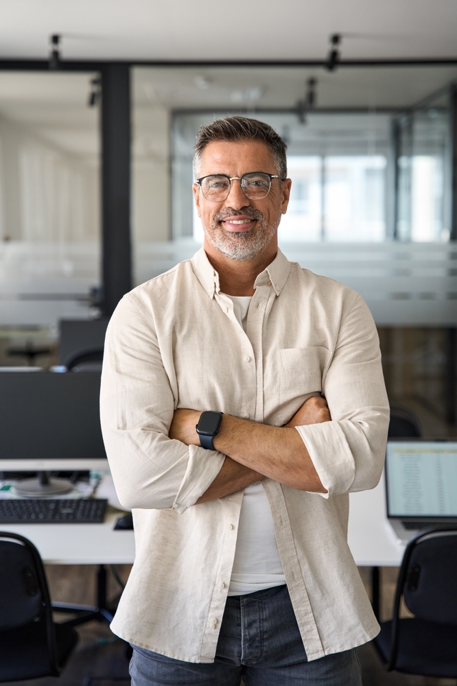 Confident middle-aged man with glasses and a casual shirt smiling with arms crossed, standing in a modern office setting