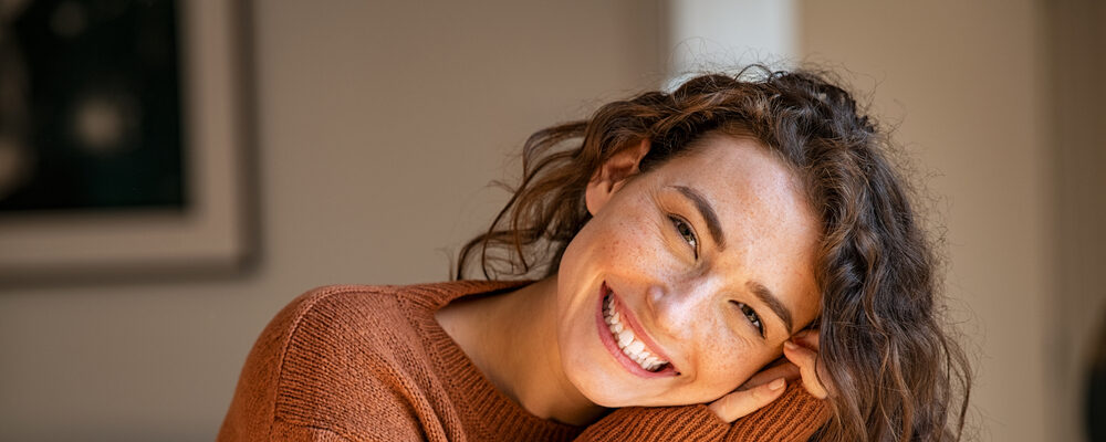 Smiling young woman with curly hair wearing a cozy brown sweater, leaning on her hands indoors.